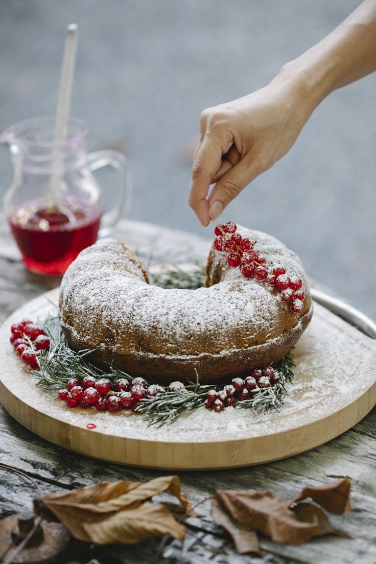 Crop anonymous female adding redcurrant in festive cake with sugar powder on round wooden board