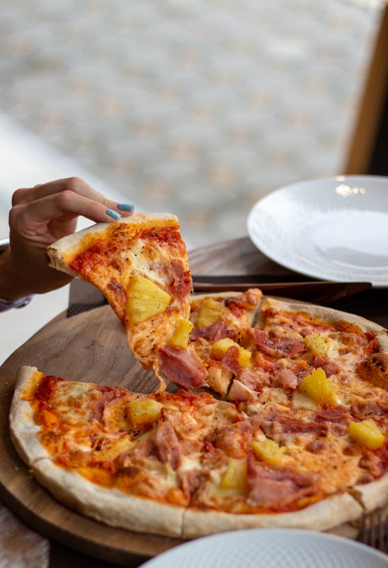 A close-up of a hand picking a slice from a freshly baked Hawaiian pizza on a wooden board.