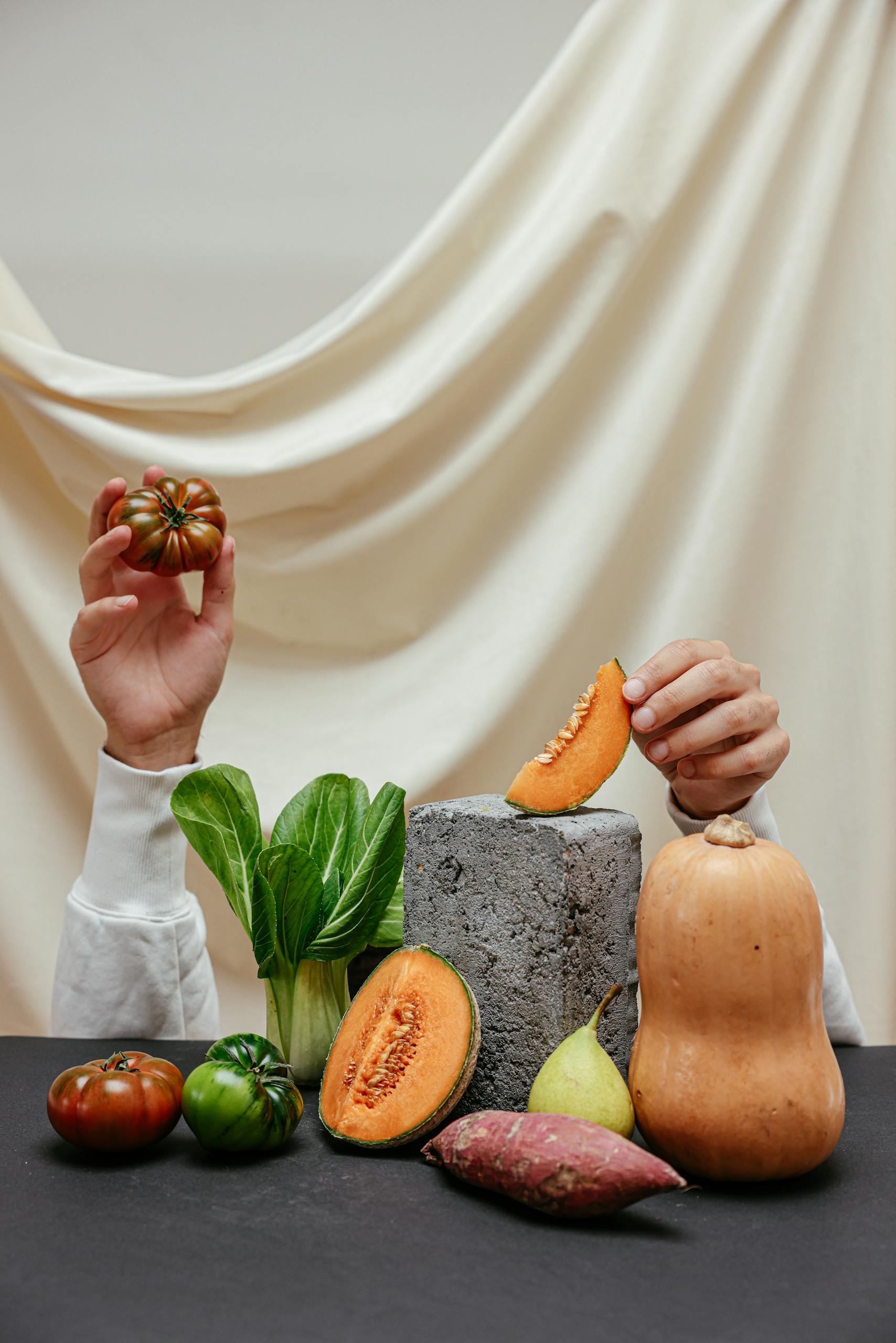 Hands holding vegetables and fruits in a creative display with a neutral backdrop.